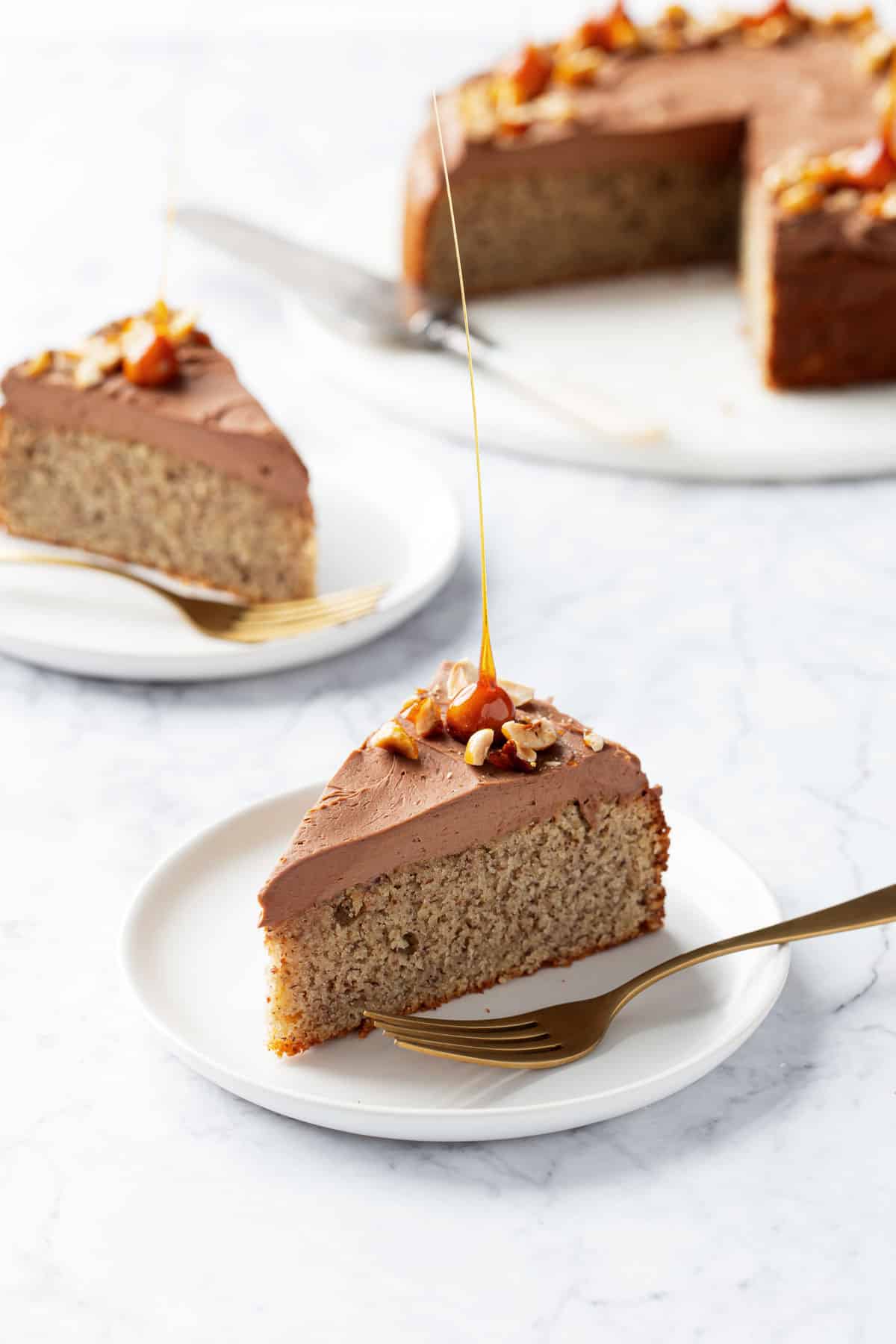 Two slices of Flourless Hazelnut Cake with Nutella Whipped Ganache on white plates with gold forks, rest of cake out of focus in the background.