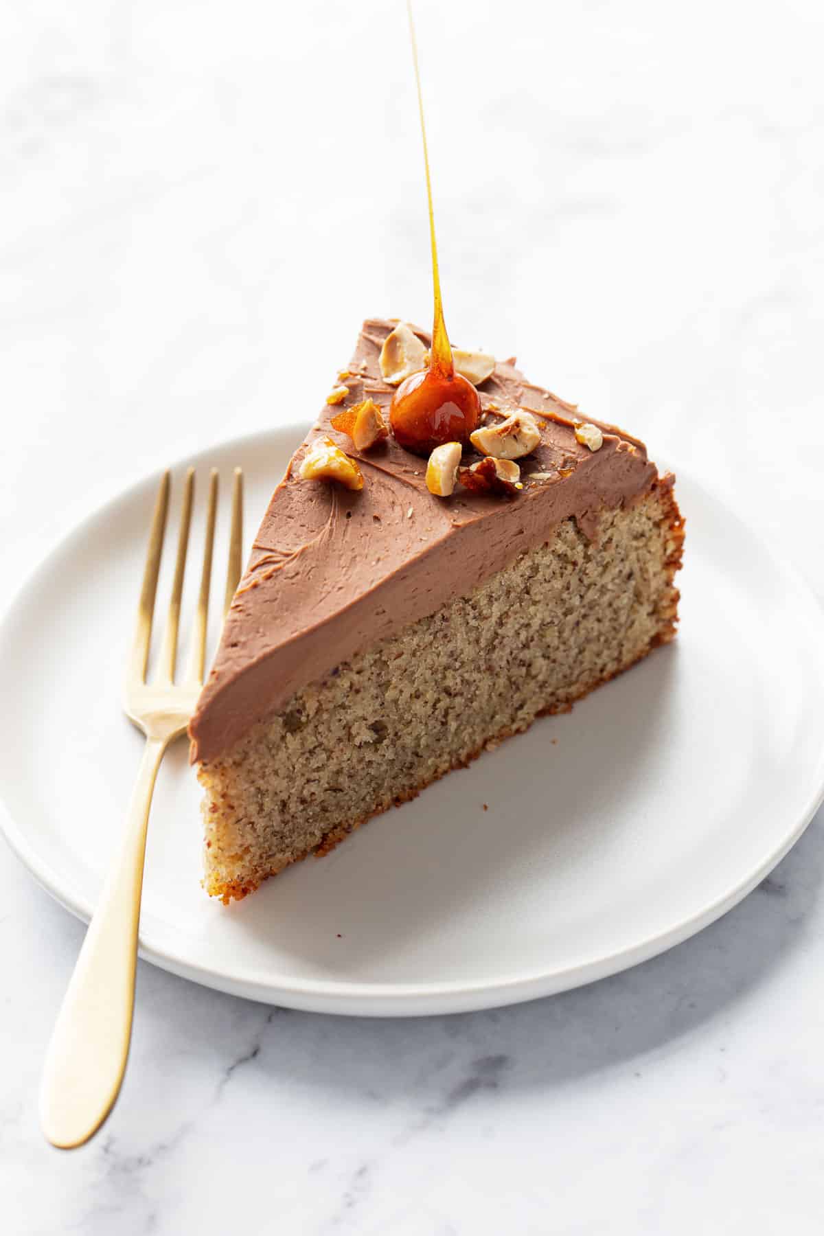 Single sharply cut slice of Flourless Hazelnut Cake, topped with a layer of Whipped Nutella Ganache and candied hazelnuts, on a white plate with gold fork.