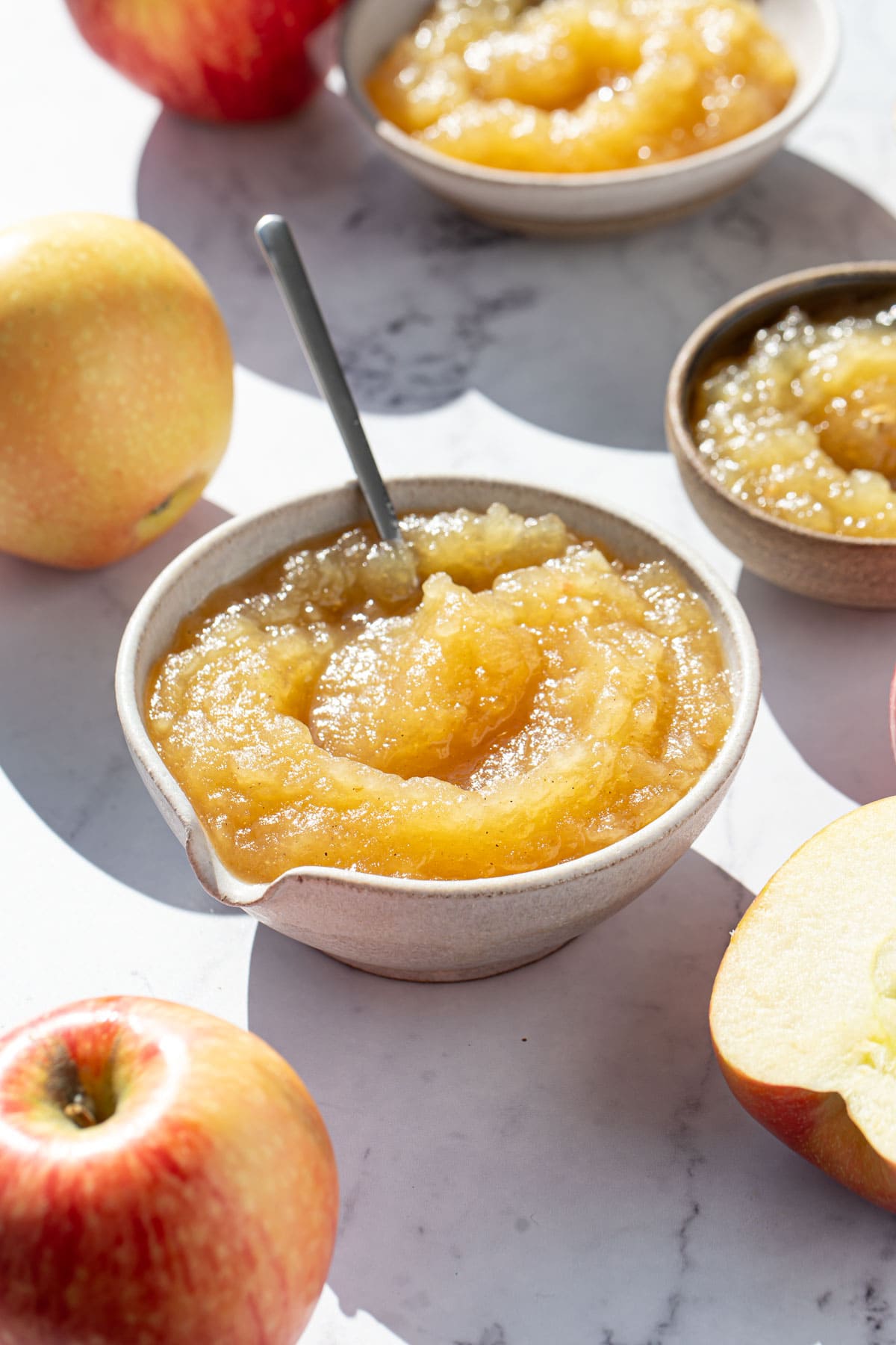 Bowl of homemade applesauce in direct sunlight, with a few smaller bowls and whole and halved apples around it.