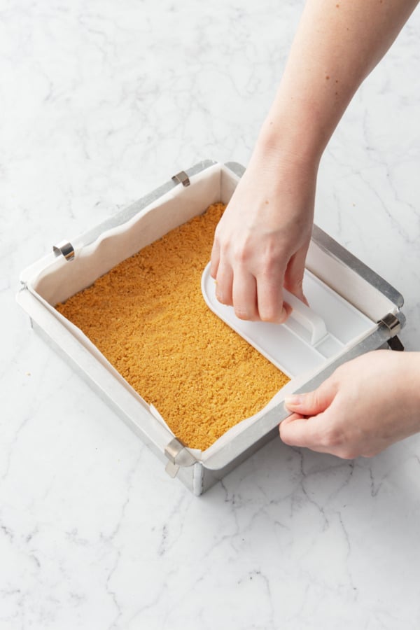 Pressing cookie crust into the bottom of a square baking pan lined with parchment paper.