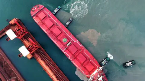 Costfoto/NurPhoto/Getty Images Aerial shot of red cargo ship with smaller ships dotted around unloading goods