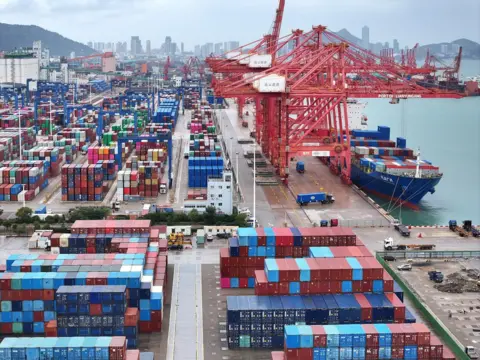 Reuters Red, blue and white containers sit stacked up on the side of a port. In the water is a cargo ship with containers being loaded on and off.