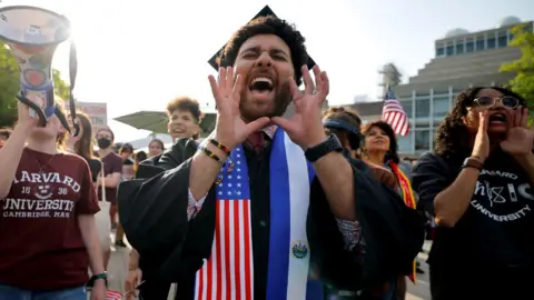 Reuters A Harvard University student chants with fellow demonstrators during a protest in support of international students at Harvard University. He is wearing a graduation cap and gown with a sash which has an American flag on it.