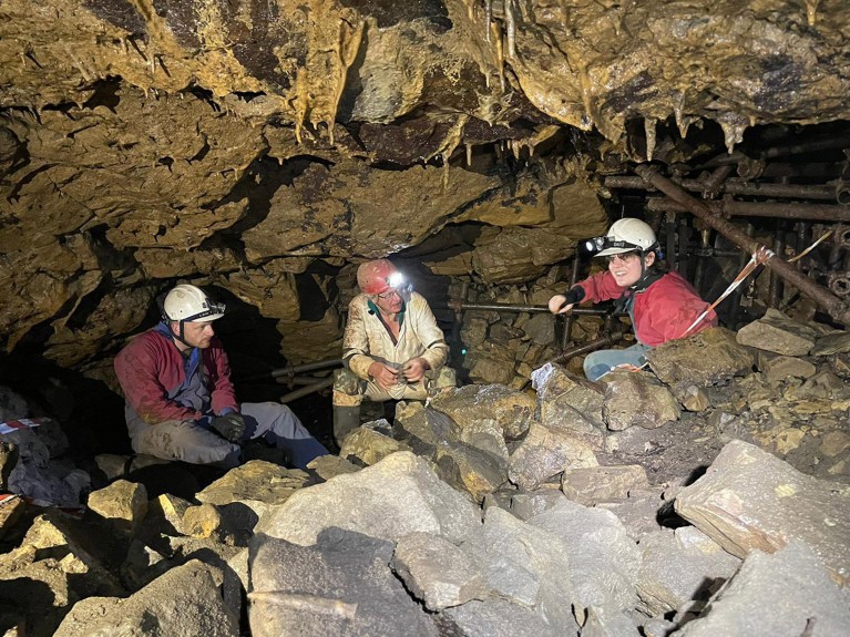 Three researchers wearing hardhats with lanterns crouched amongst rocks in the Bone Idle Chamber of Fishmongers Swallet.