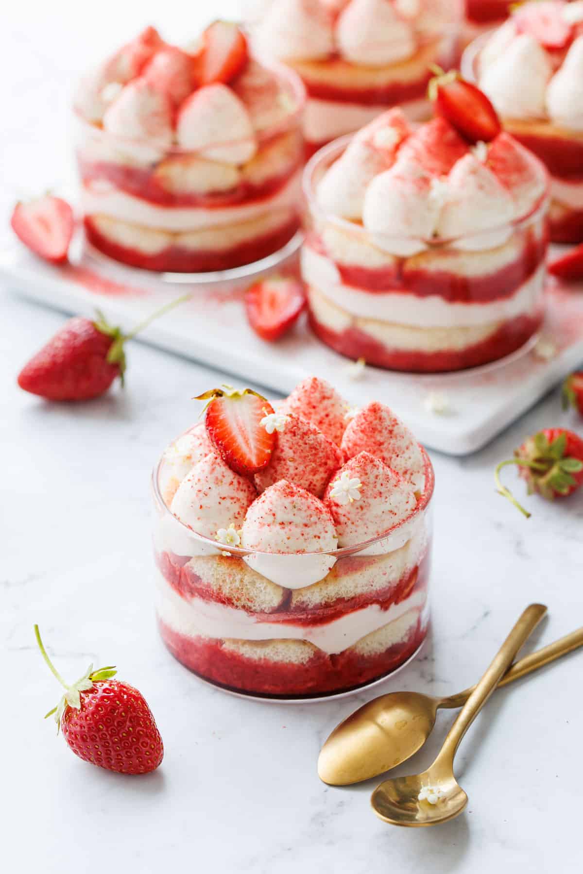 Strawberry Elderflower Tiramisu in individual clear ramekins, on a white marble background with two gold spoons and strawberries.