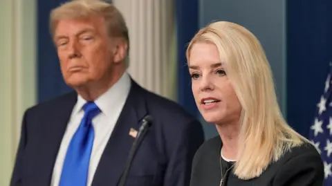 Reuters US Attorney General Pam Bondi, a woman with short blonde hair wearing a black blouse, speaks in front of a US flag, with US President Donald Trump behind her wearing a dark blue jacket, white shirt and bright blue tie.