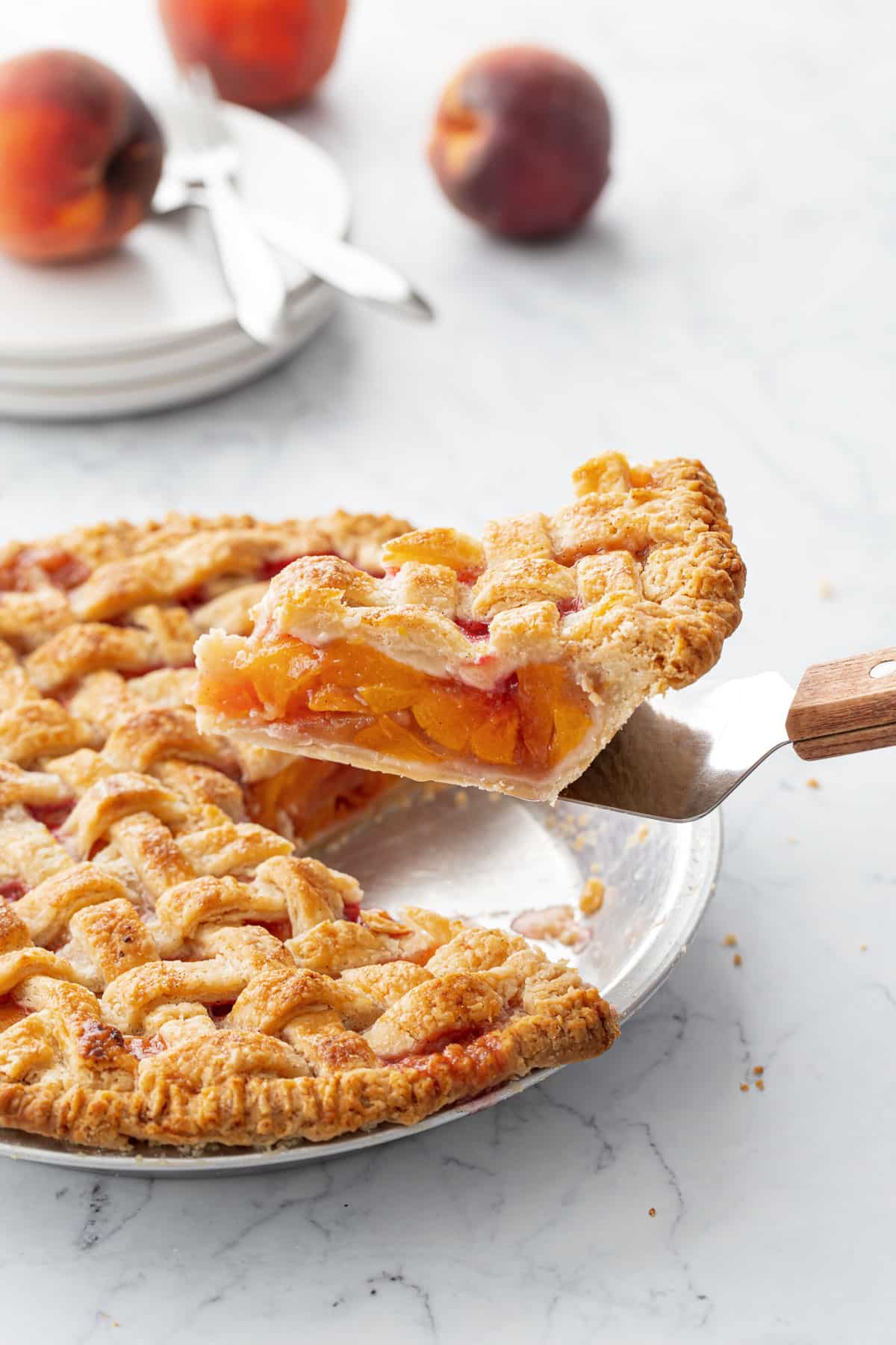 Slice of Vanilla Bean Peach Pie being lifted from the pan, with a stack of plates and fresh peaches in the background.