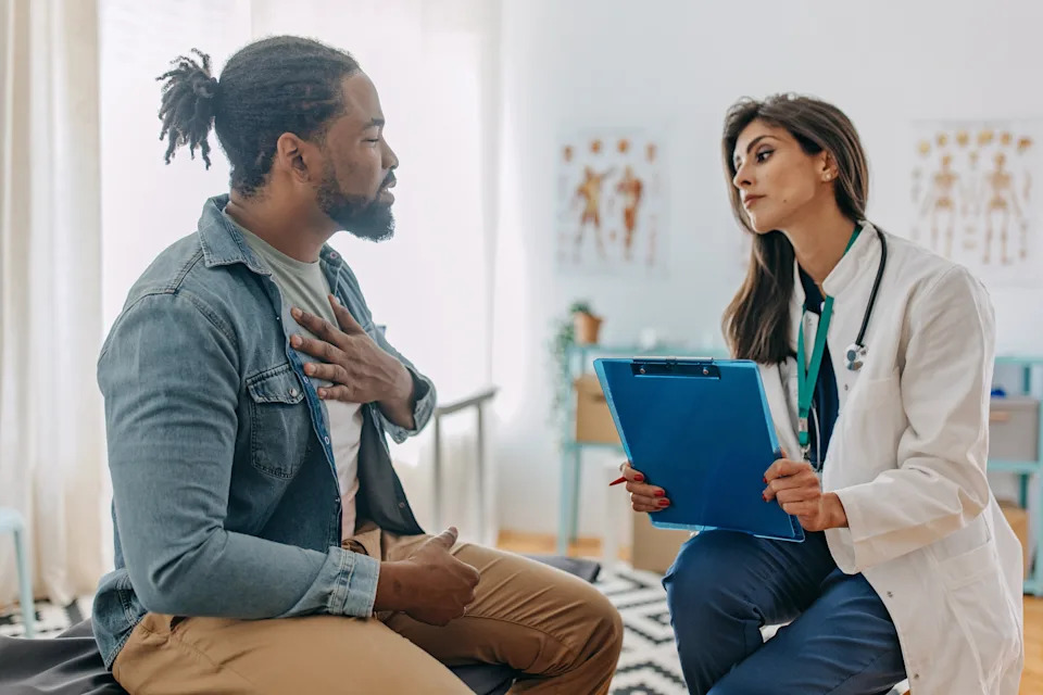 A man consults with a doctor in an office; she holds a clipboard while attentively listening to him