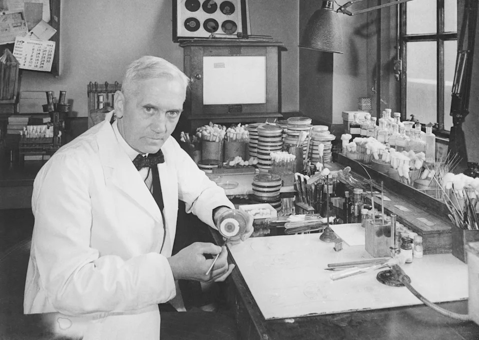 An older scientist in a lab coat works at a cluttered lab bench with petri dishes and tools