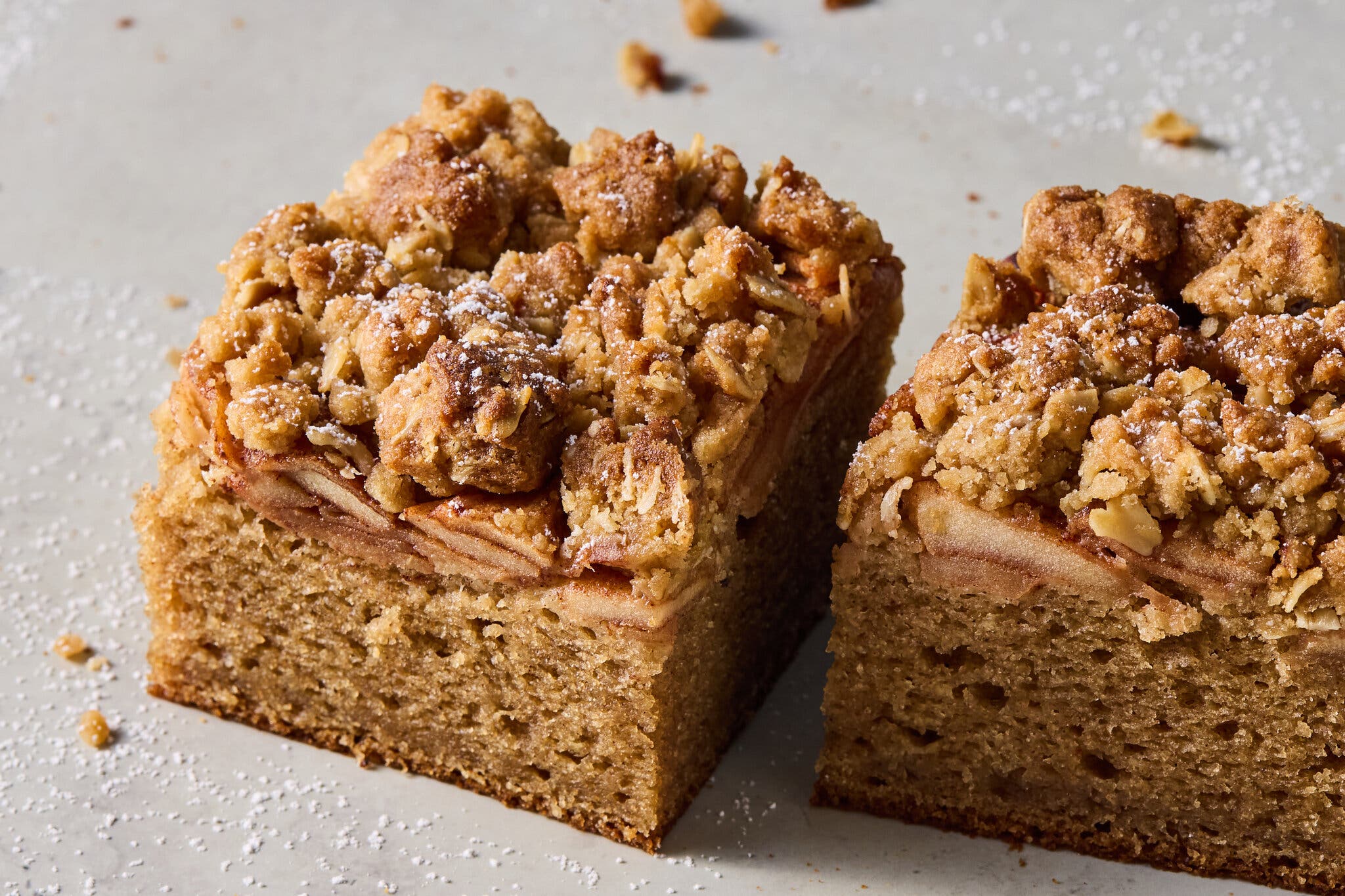 Two square slices of crumb cake are dusted with powdered sugar and sit on a white background.