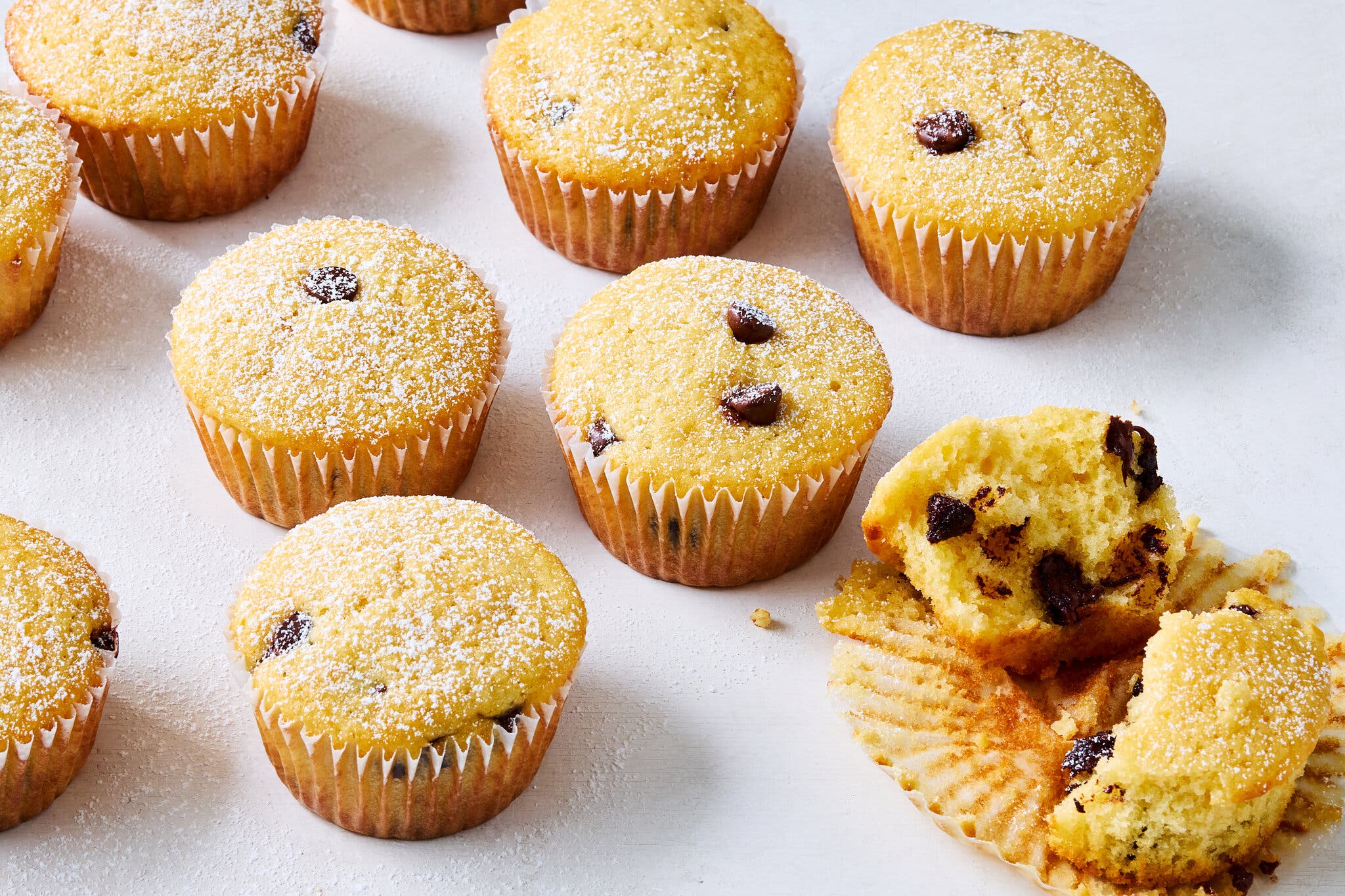 Chocolate chip muffins, dusted with powdered sugar, sit on a white surface.