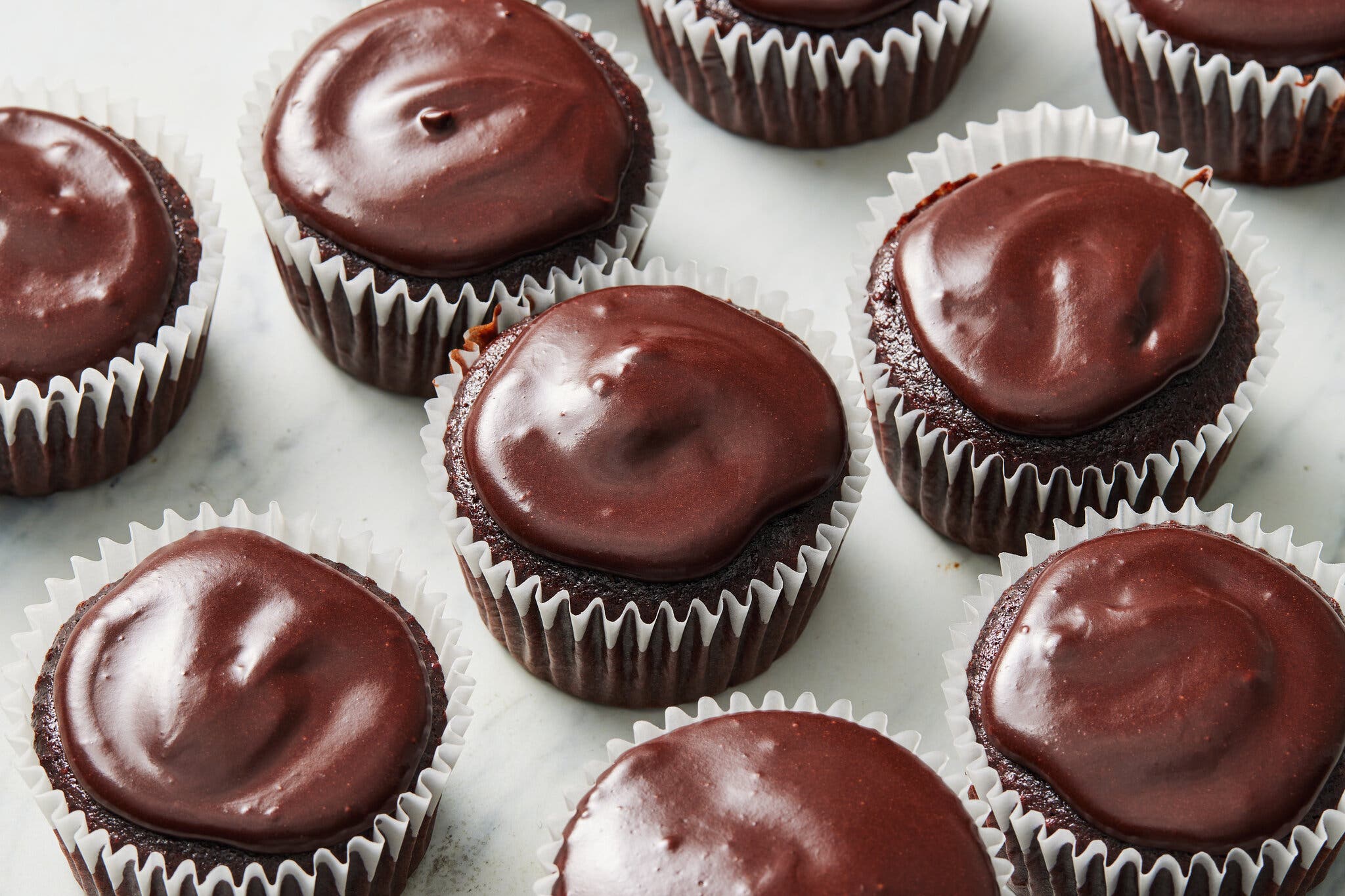 Chocolate cupcakes with dark, glossy frosting are arranged on a white marble surface.