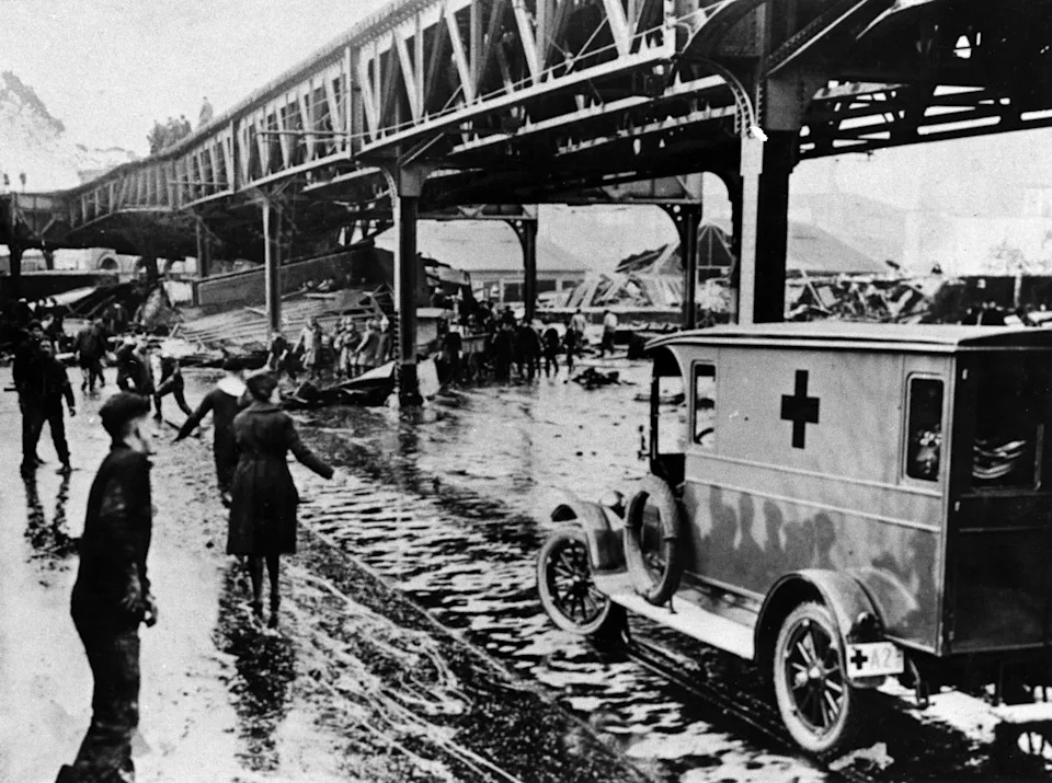 Historical scene of a disaster with rescue workers, a Red Cross vehicle, and bystanders near a damaged elevated railway structure