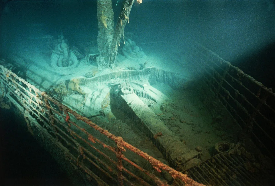 Underwater view of the Titanic's bow on the ocean floor, showing the ship's railings and decks, partially covered in silt and marine growth
