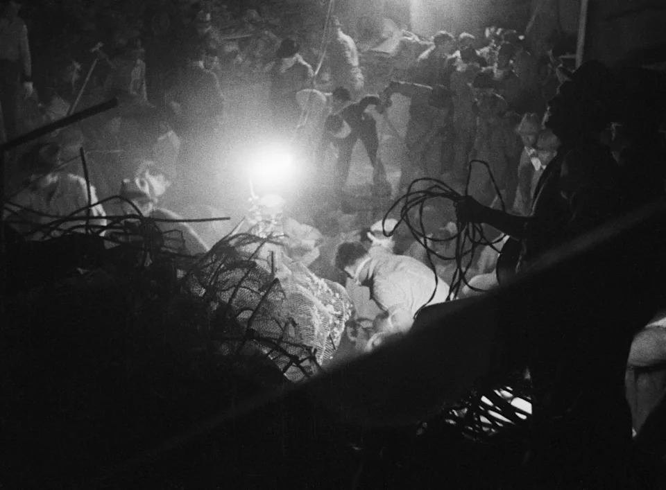 Rescue workers and onlookers gather around a disaster site at night, with bright lights illuminating their work amidst debris and tangled wires
