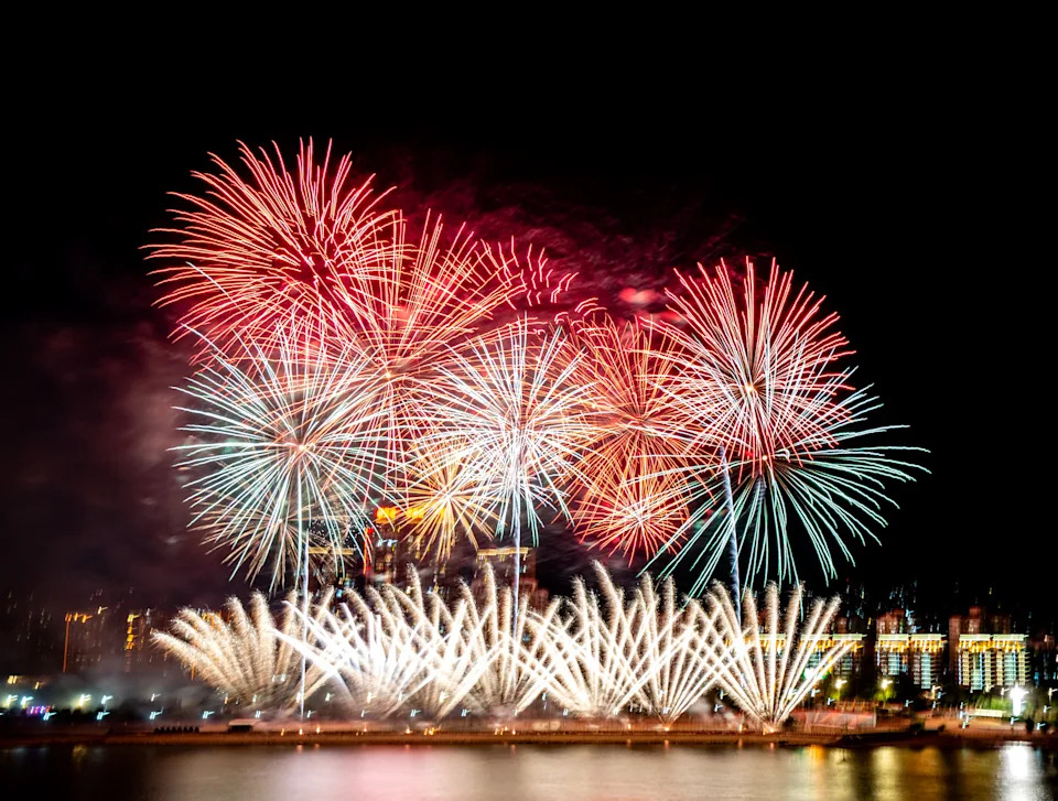 Fireworks burst in the night sky above a cityscape, illuminating buildings and reflecting over water