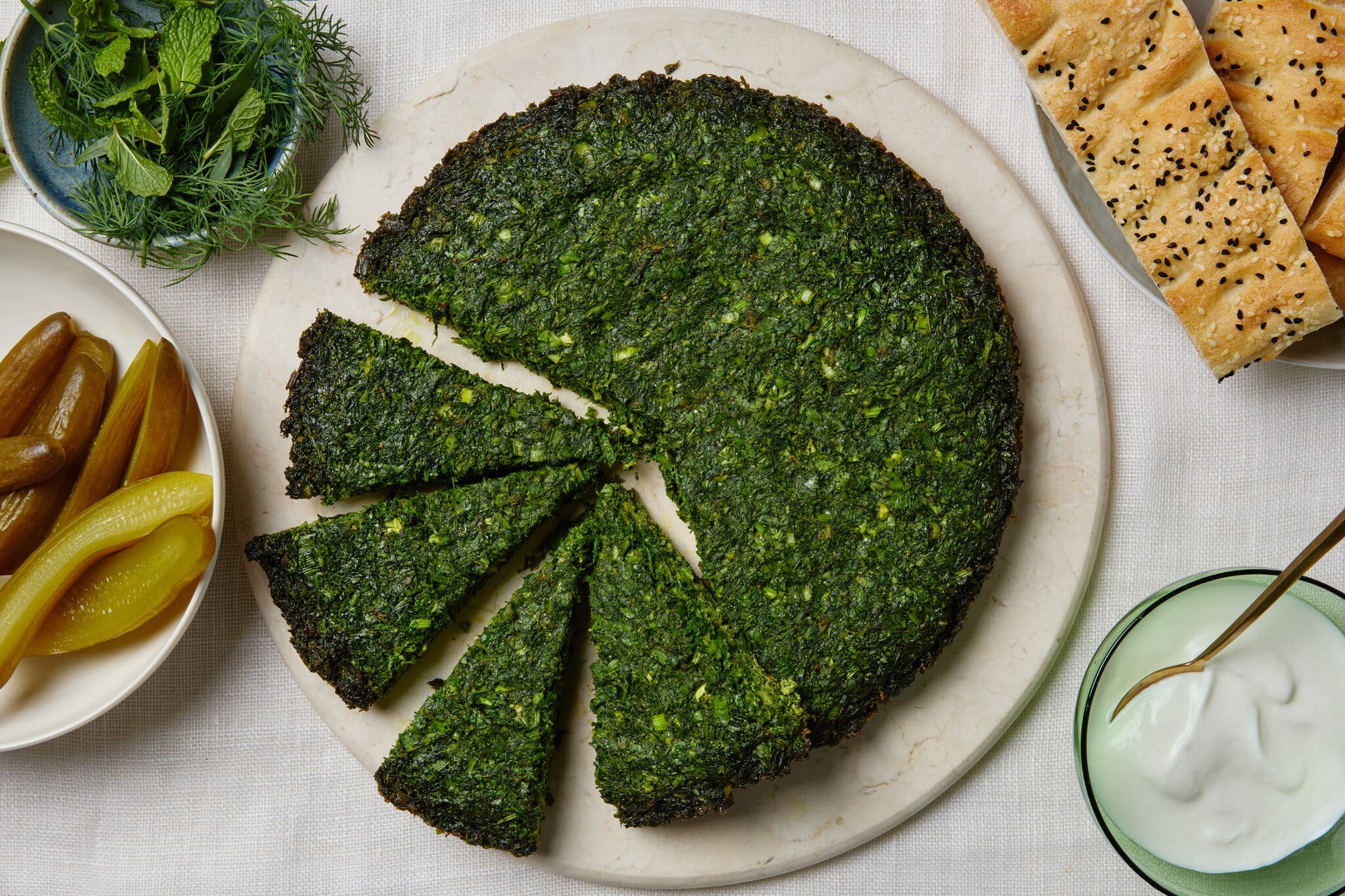 An overhead image of a bright green frittata surrounded by flatbread, herbs, pickles and yogurt.