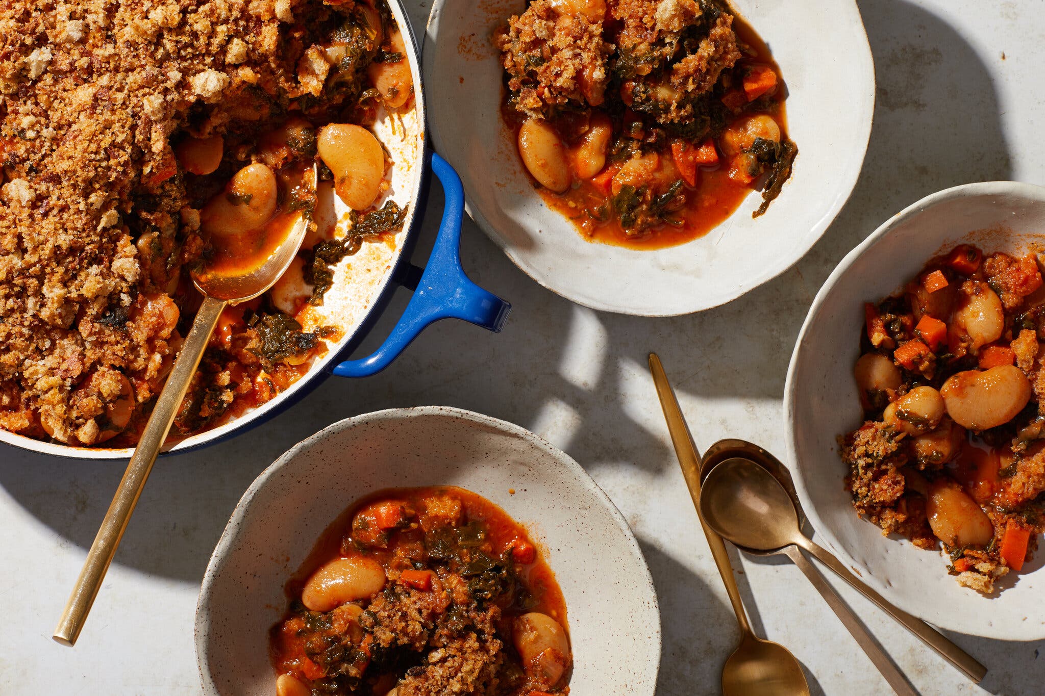 An overhead image of a large pot filled with beans and kale, and topped with bread crumbs. A few servings surround the pot.